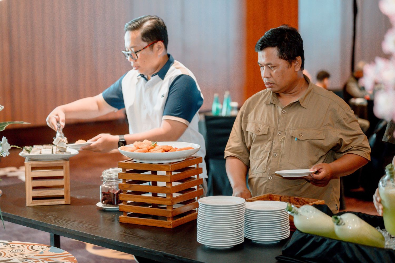 Two men serving themselves at a buffet with stacked white plates and wooden food display stand.