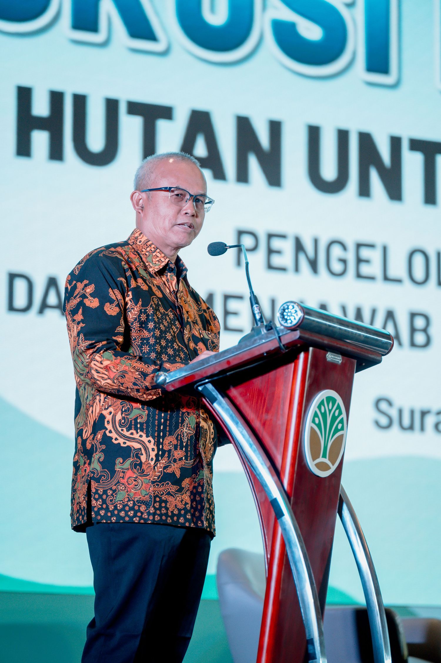 Indonesian official in batik shirt speaks at podium during Hutan forest management discussion event in Surabaya.