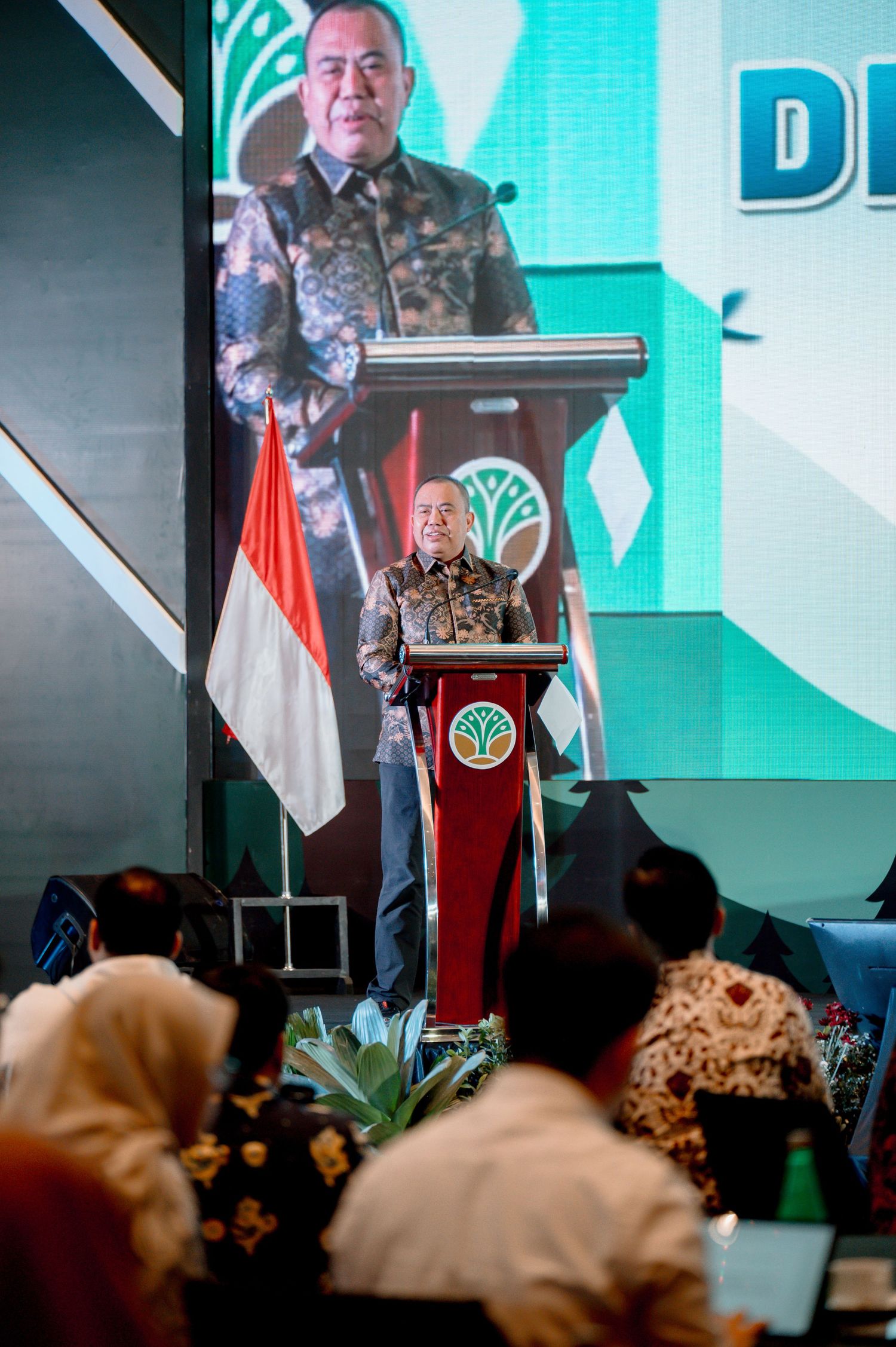 Indonesian official in batik shirt delivers speech at podium with national flag and green logo backdrop.