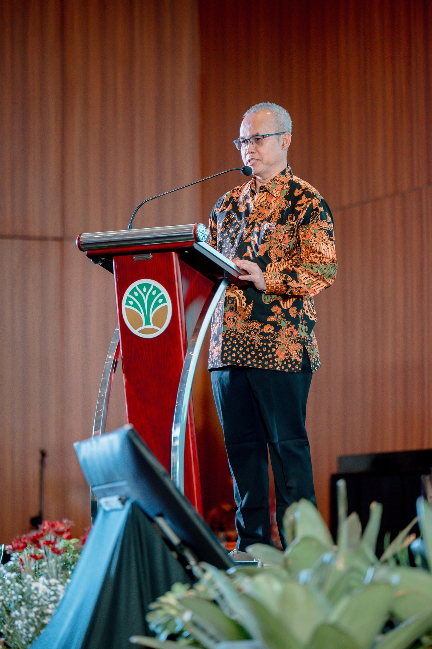 Man in batik shirt speaking at a red podium with a green tree logo at a formal Indonesian event.