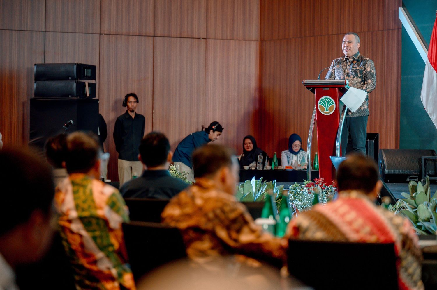 Indonesian official delivers speech at podium during formal government conference with attendees in traditional batik attire.