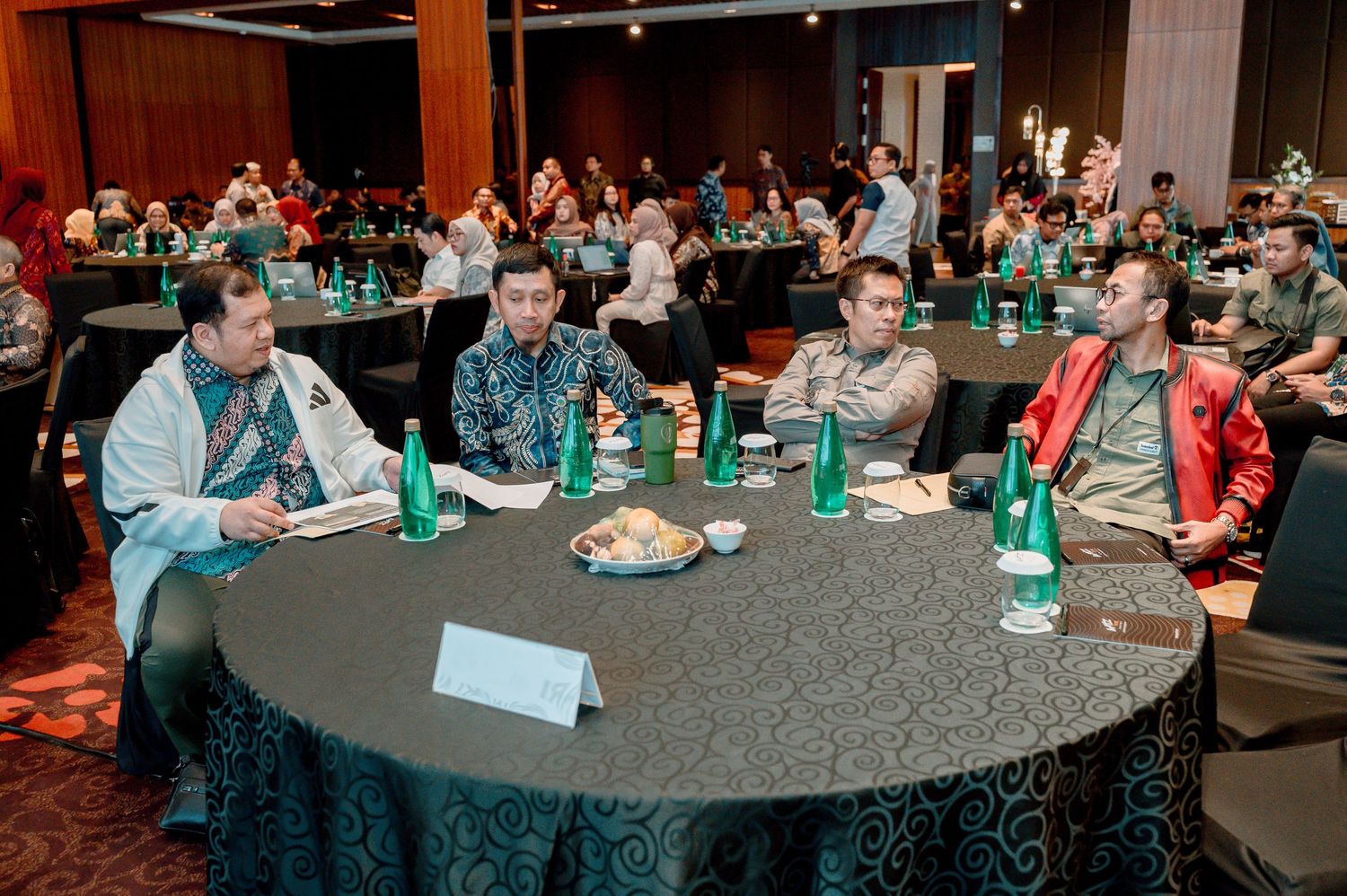 Four men in formal attire sit at a round table during a corporate conference event in a large banquet hall.