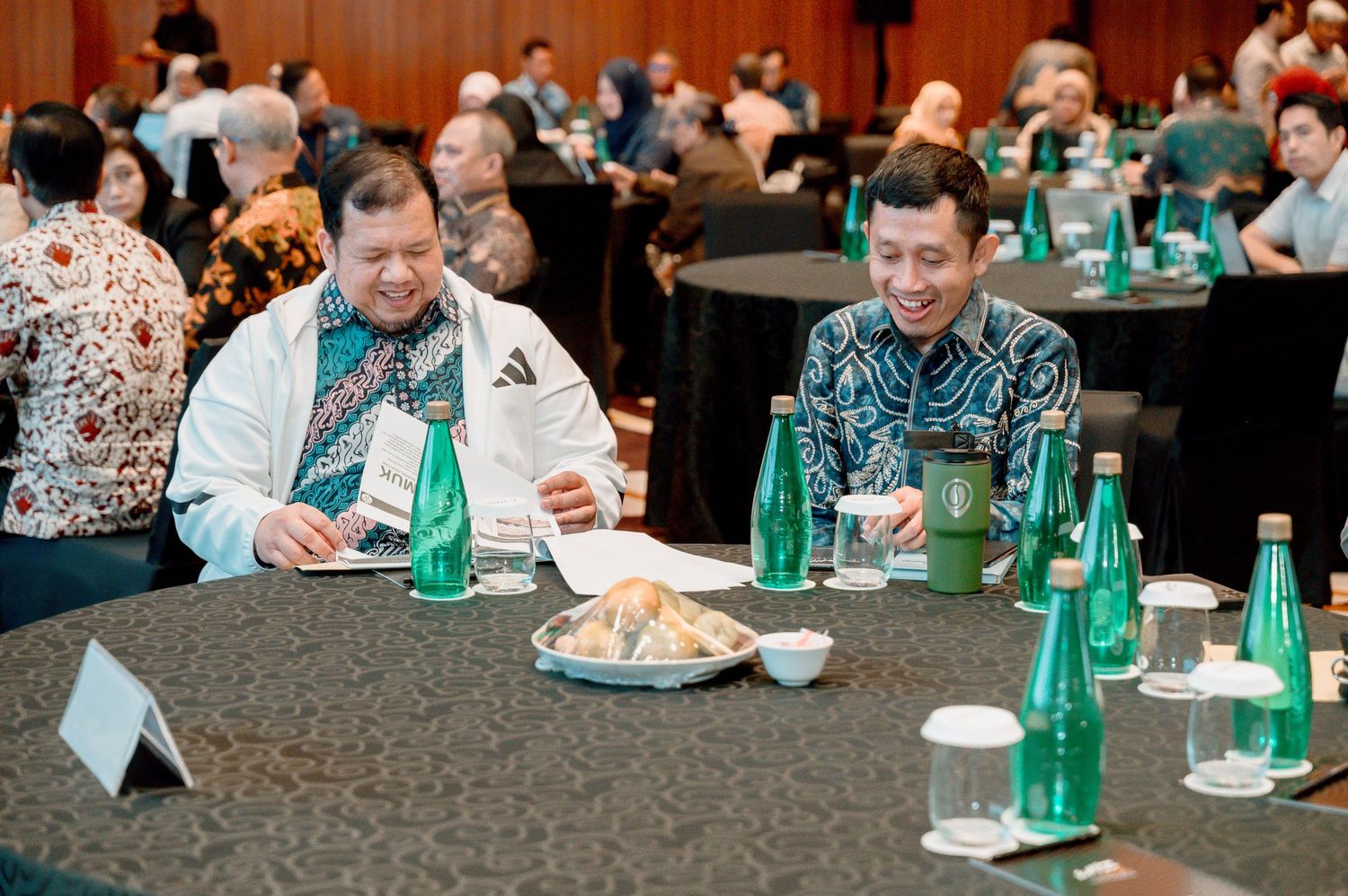 Two men in batik shirts smile at a formal dinner event with green water bottles on a dark round table.