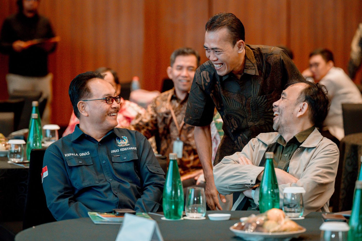 Three men in formal attire share a laugh at a conference table with green water bottles.