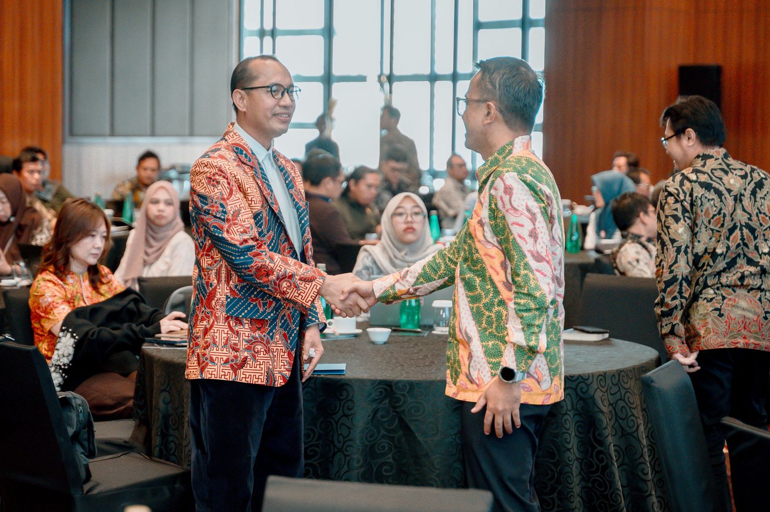 Two men in batik shirts shake hands at a formal Indonesian business conference event.