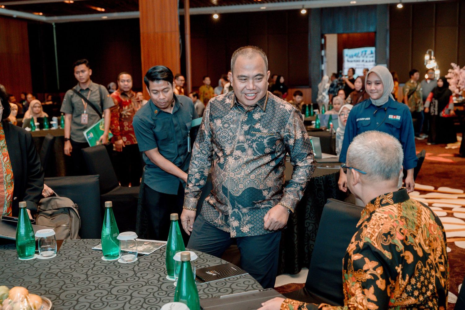 Indonesian officials in batik attire networking at a formal conference event in a modern meeting hall.