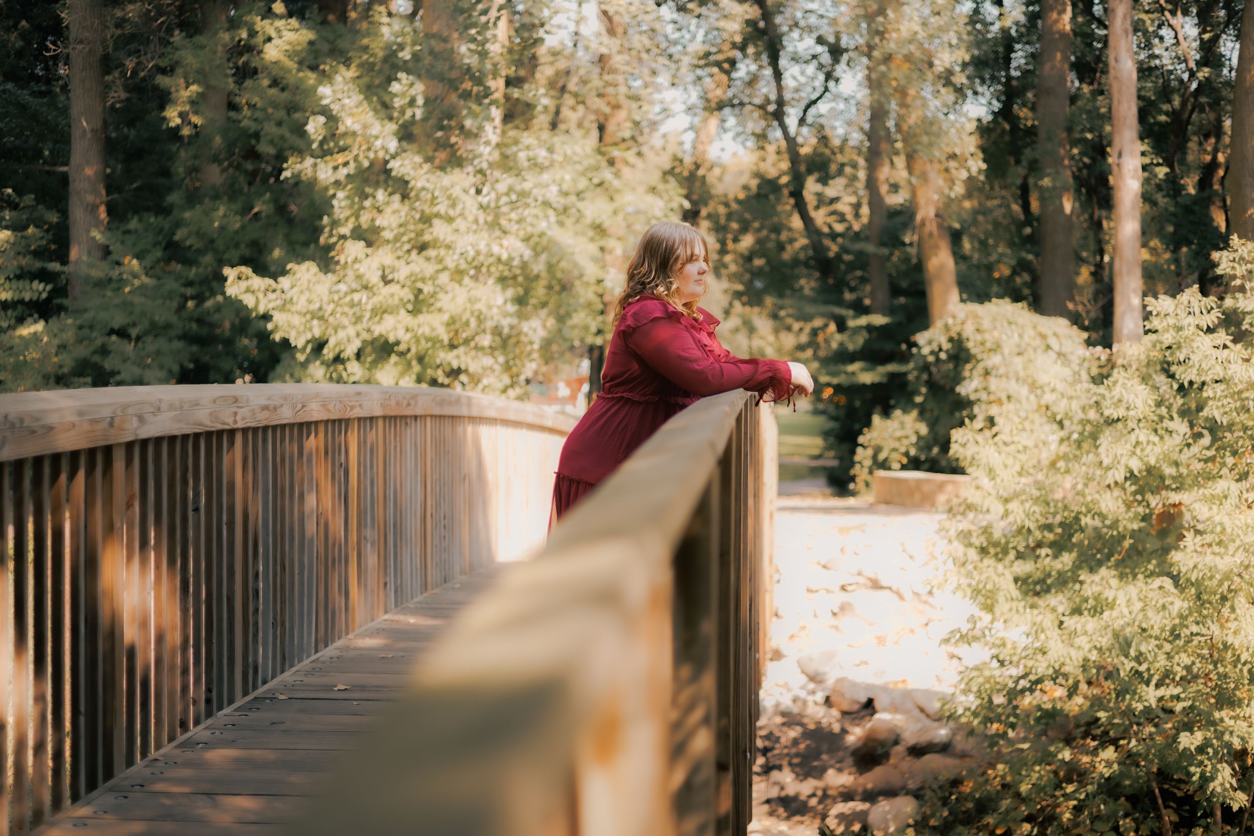 Woman in red dress leaning on wooden bridge railing surrounded by lush green trees in a serene park setting.