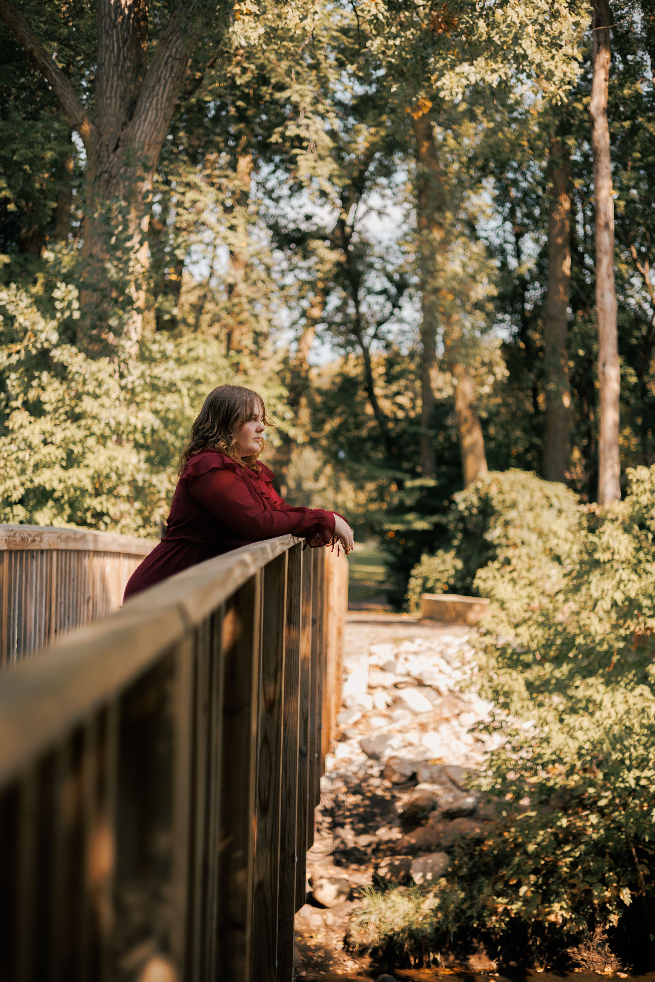 Woman in burgundy jacket leaning on wooden bridge railing, gazing into lush green forest park surroundings.
