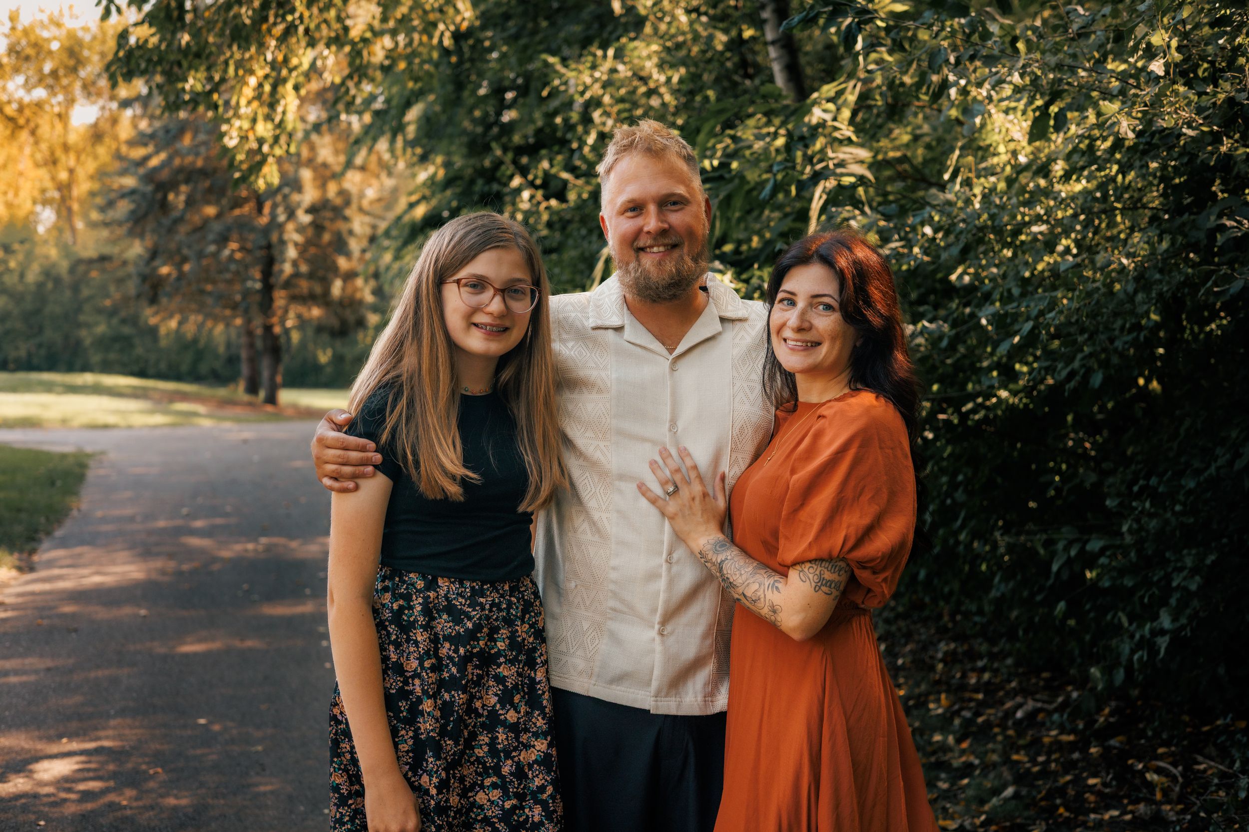 A family of three smiling outdoors in a lush green park during golden hour.