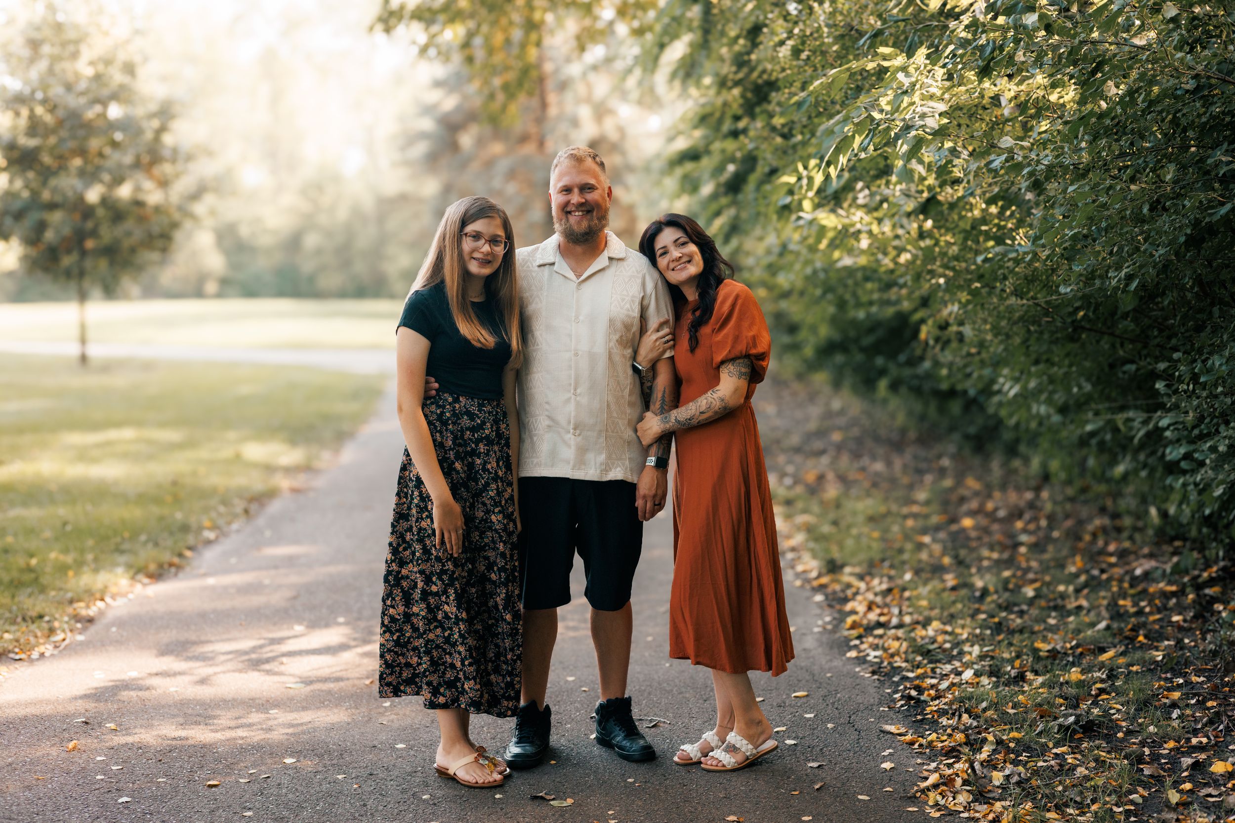 Three people smiling together on a park path surrounded by lush green trees on a sunny day.