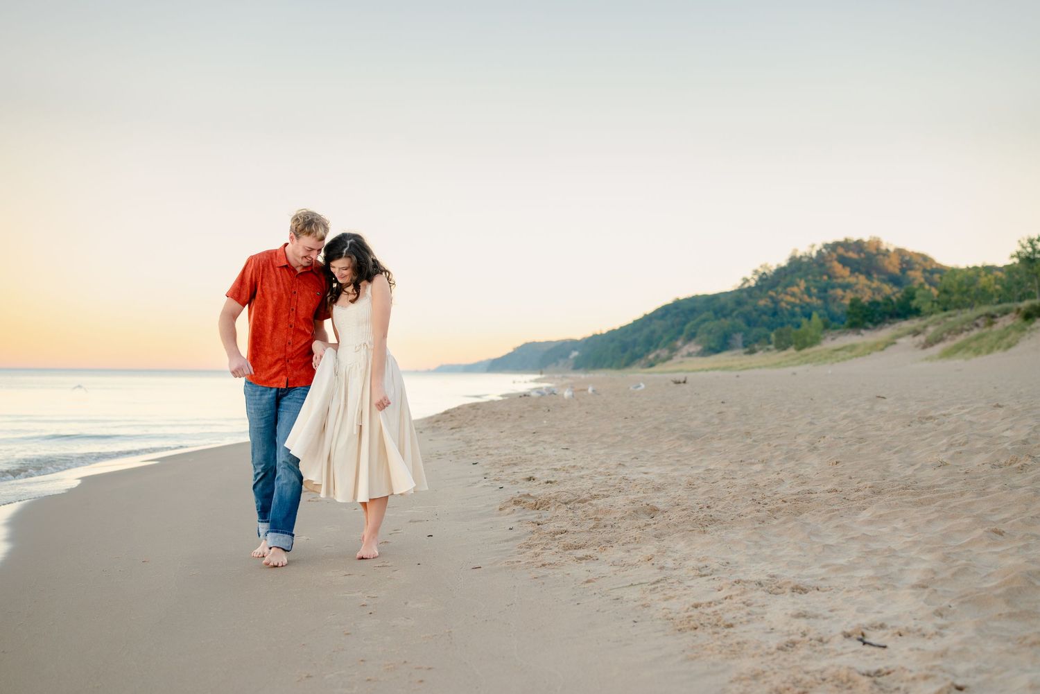 A joyful couple on a beach at sunset, man in red shirt lifting woman in white dress near calm ocean waters.