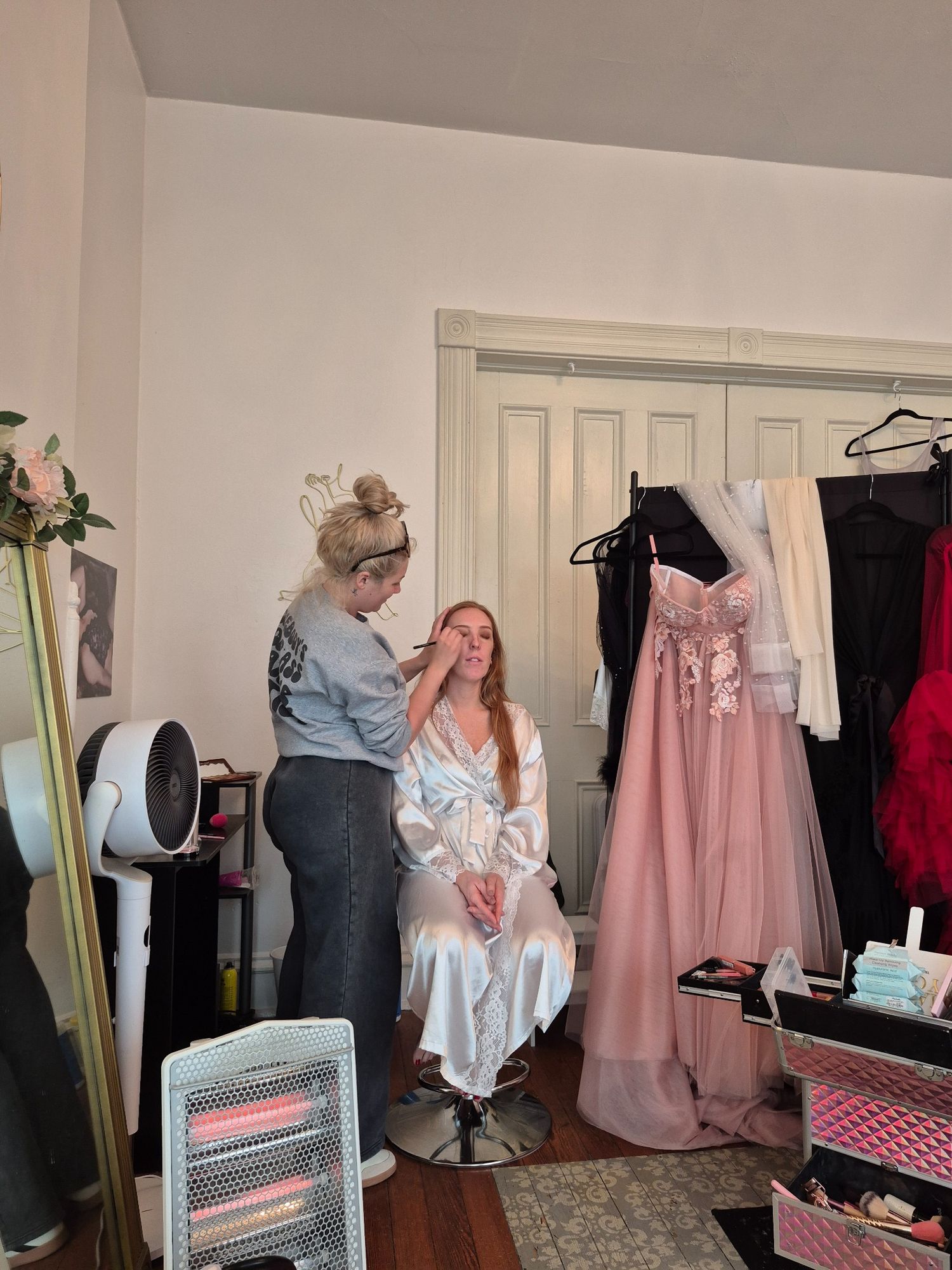 A makeup artist applies makeup to a seated woman in a white robe near a pink prom dress in a salon.
