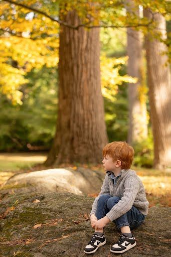 Red-haired boy sitting on a rock in an autumn forest park, wearing a gray sweater and jeans.
