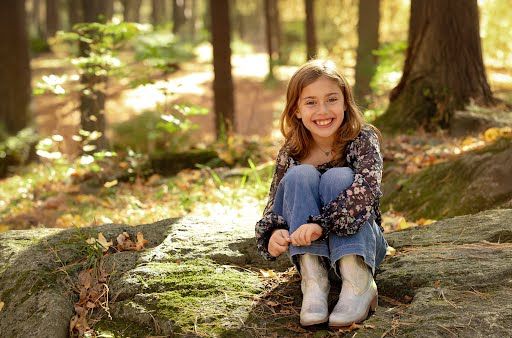 Smiling young girl sitting on a mossy rock in a sunlit forest, wearing jeans and boots.
