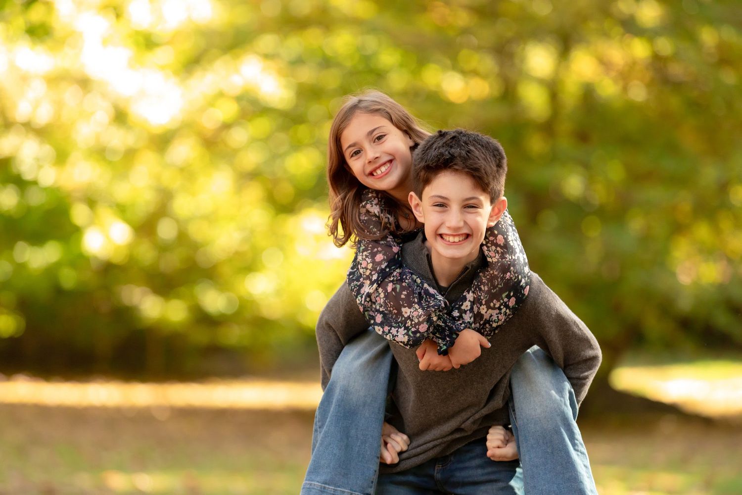 Two happy children pose outdoors in autumn, with a boy giving a smiling girl a piggyback ride among golden trees.