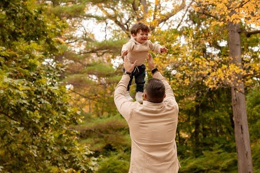 Father joyfully lifting his laughing toddler in the air outdoors in an autumn park with colorful foliage.