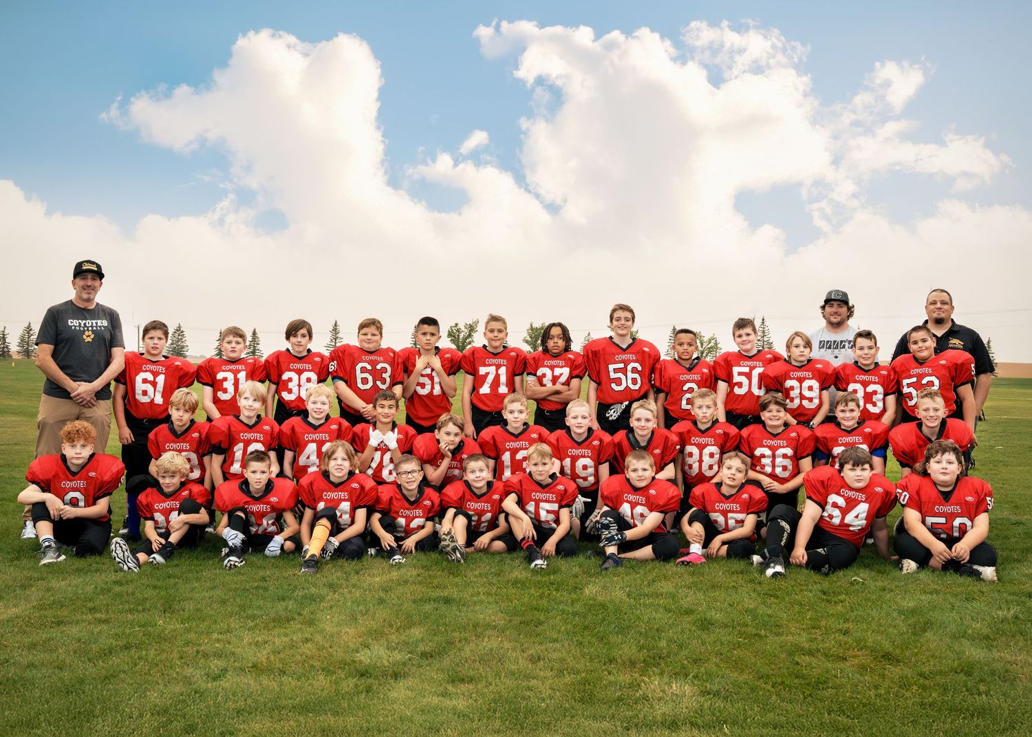 Youth football team poses for group photo outdoors in dark uniforms, coaches and players lined up on grass field.