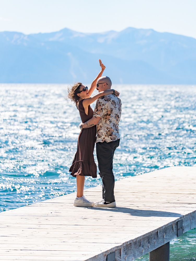 Couple dancing on a wooden pier with sparkling blue lake and mountain backdrop on a sunny day.