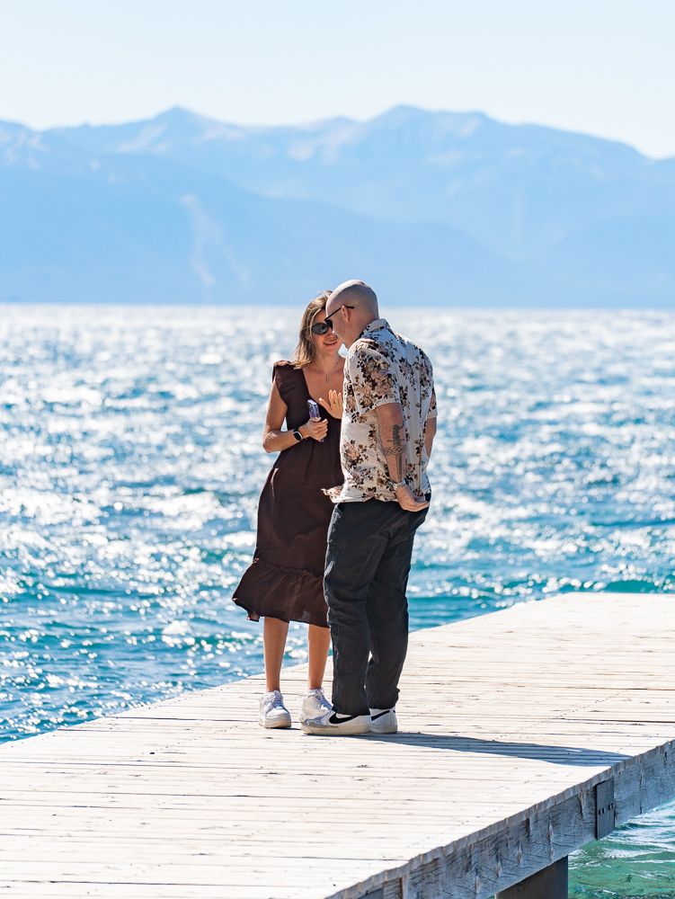 Couple kissing on a wooden dock with sparkling blue lake and mountain backdrop on a sunny day.