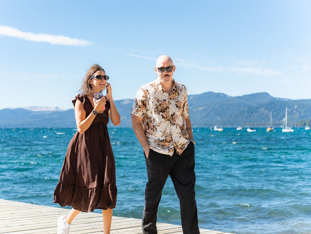 A woman in a brown dress and a bald man in a floral shirt stand on a dock by a blue lake.
