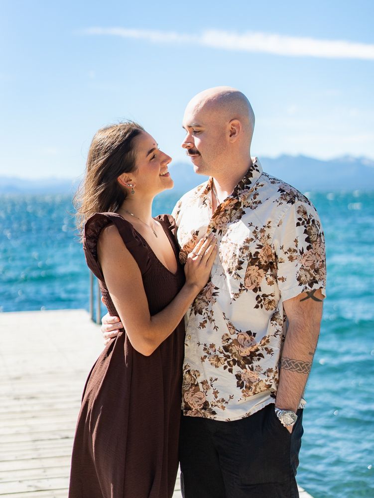 Couple smiling at each other on a lake dock, woman in brown dress, man in floral shirt, blue water background.