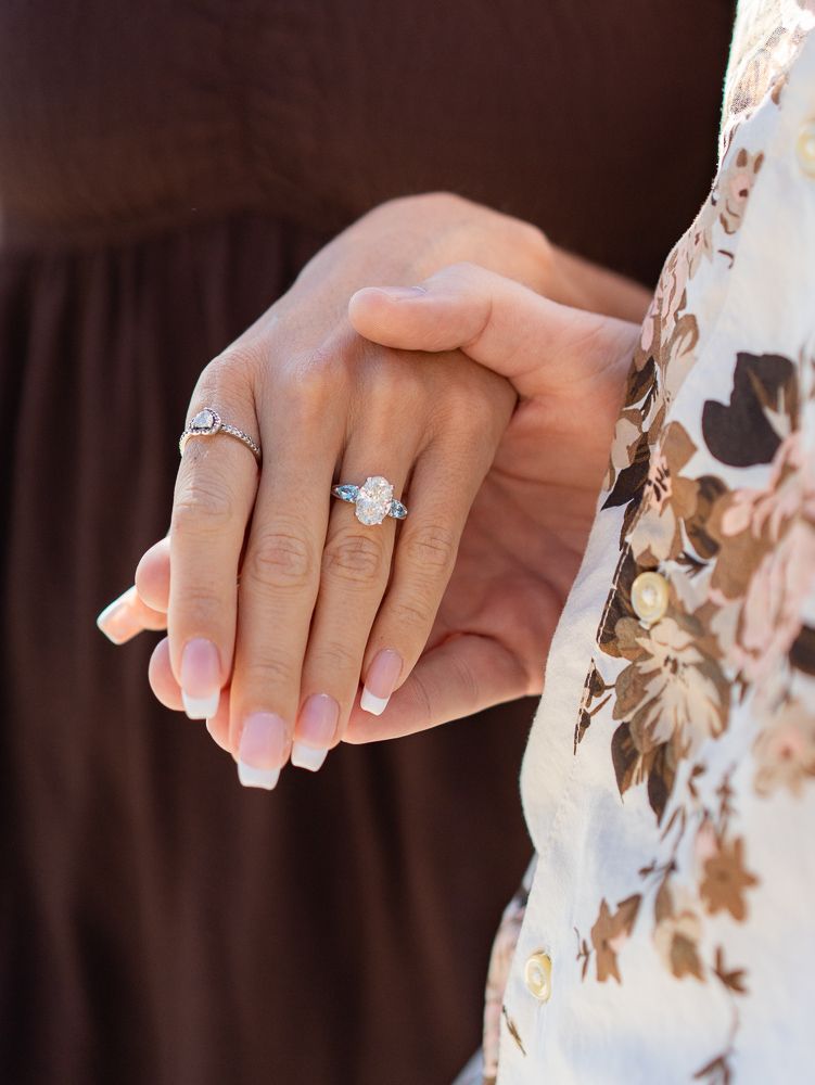 Close-up of two hands holding, showcasing a stunning oval diamond halo engagement ring with French manicured nails.