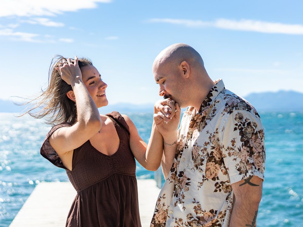 A couple laughs together on a sunny dock by blue water, woman in brown dress, man in floral shirt.