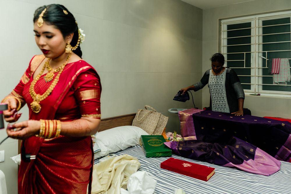 Indian bride in red saree with gold jewelry prepares for wedding ceremony while attendant irons clothes on bed.