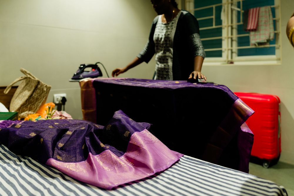 Woman ironing purple silk saree on bed with red suitcase in background.