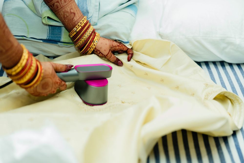 Bride with henna-adorned hands and gold bangles steaming a cream wedding garment on a striped bed.