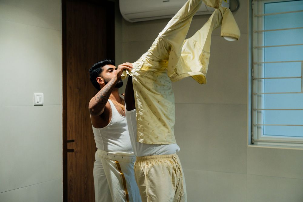 A man in a white tank top holds up a yellow traditional outfit on a hanger in a modern room.