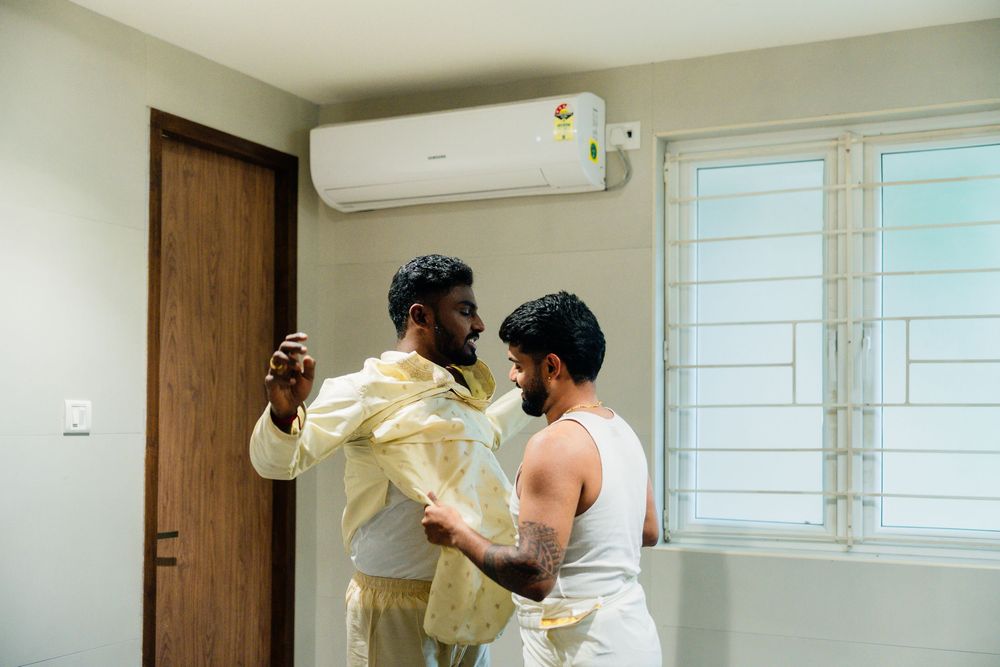 Two men help each other dress in traditional Indian wedding attire in a modern room with air conditioning.