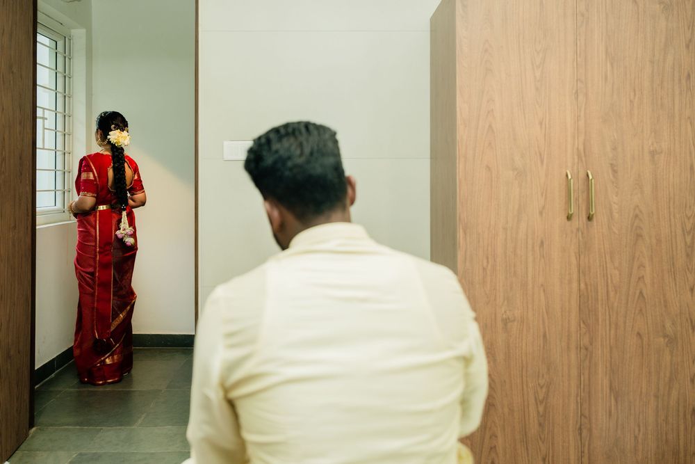 Groom in white sherwani watches bride in red saree standing before mirror during Indian wedding preparation.