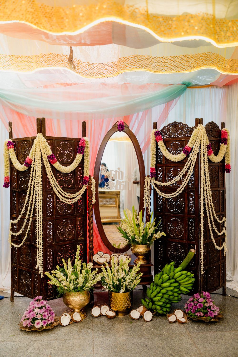 Traditional Indian wedding decor with carved wooden screens, jasmine garlands, mirror, banana leaves, and colorful flowers under draped fabr