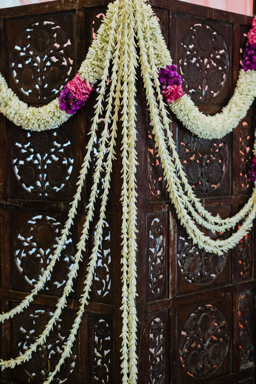 Ornate carved wooden screen adorned with white jasmine garlands and purple flower accents for a traditional Indian wedding.