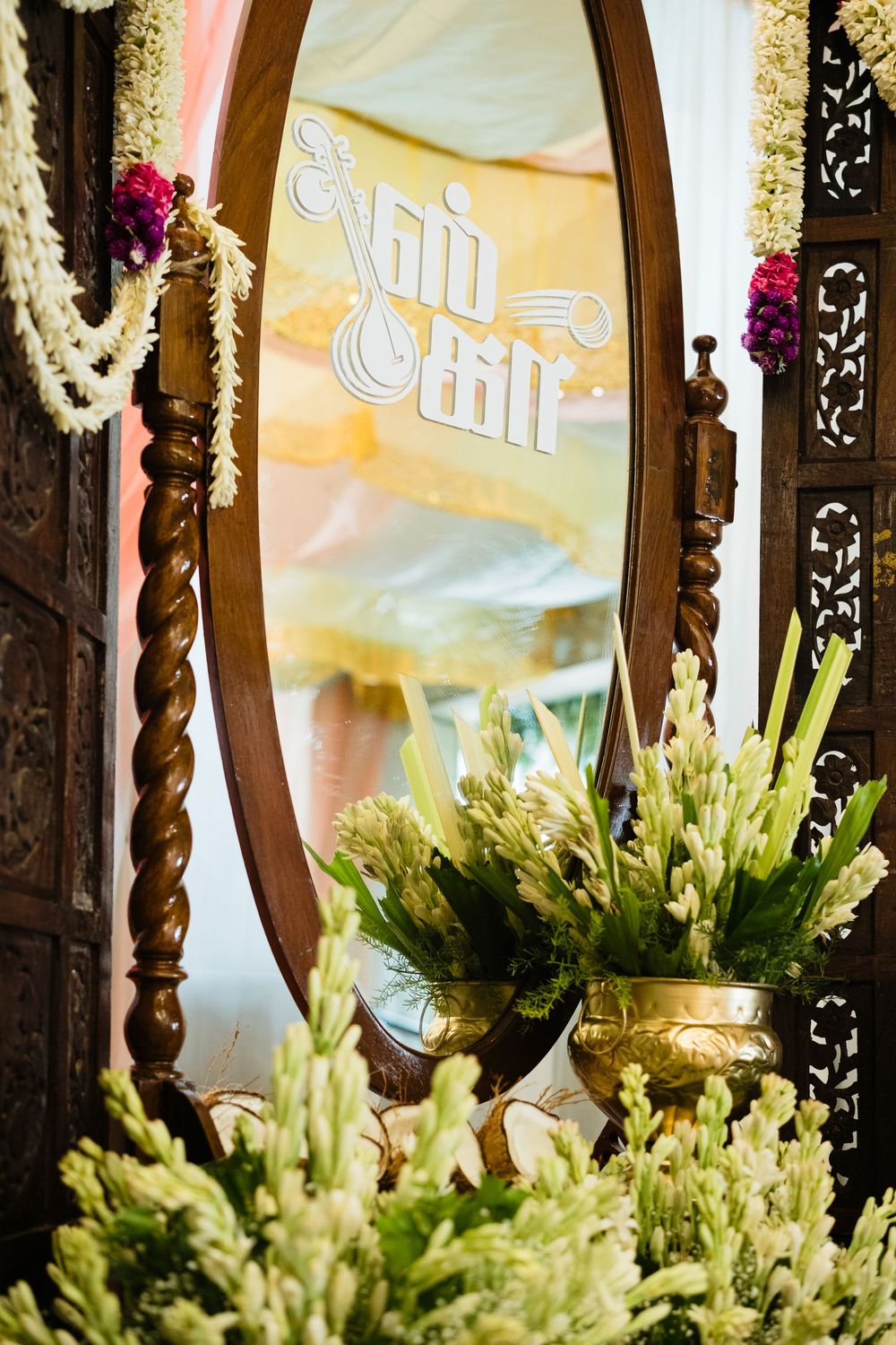 Ornate wooden mirror with Tamil text reflecting floral decorations and white tuberose arrangements at an Indian wedding ceremony.