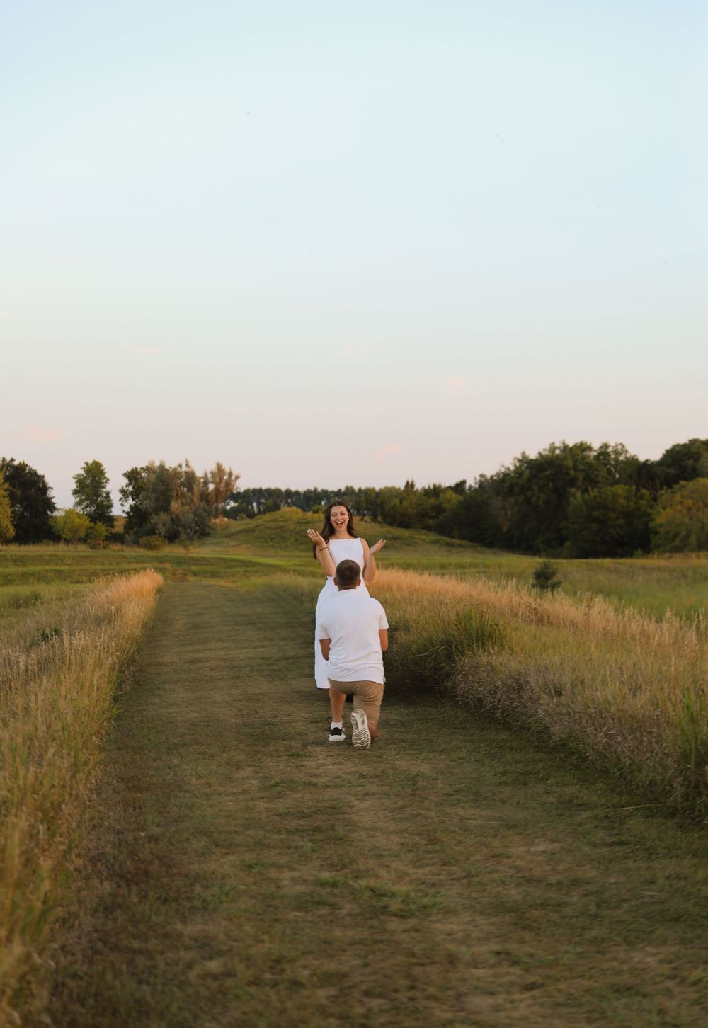 A man kneeling to propose to a woman on a scenic grass path surrounded by fields at golden hour.