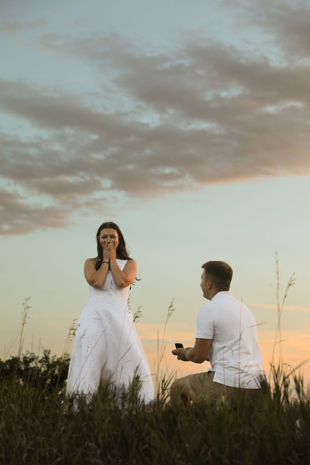 Man proposes on one knee to surprised woman in white dress at sunset in a grassy field.