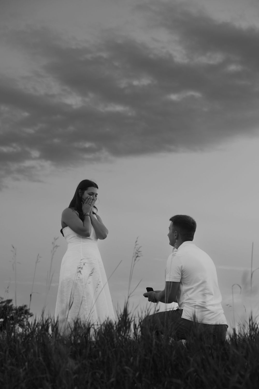 Black and white photo of a man on one knee proposing to a woman in a white dress outdoors at dusk.