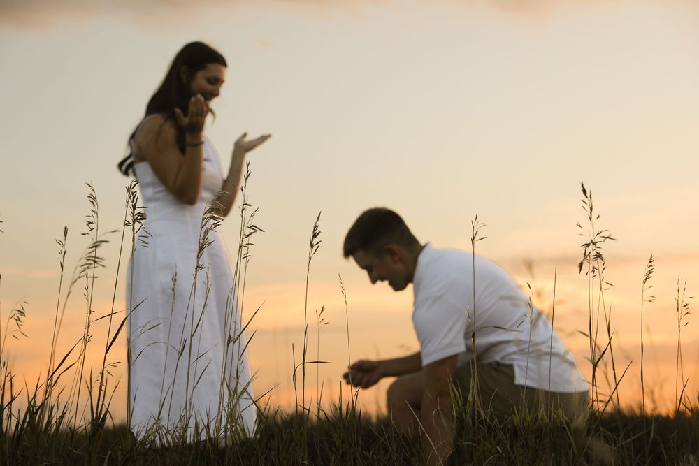 Man proposing to woman in white dress at sunset in a grassy field, romantic outdoor marriage proposal scene.