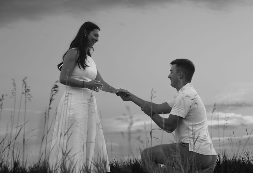 Black and white photo of a man on one knee proposing to a woman in a field at dusk.