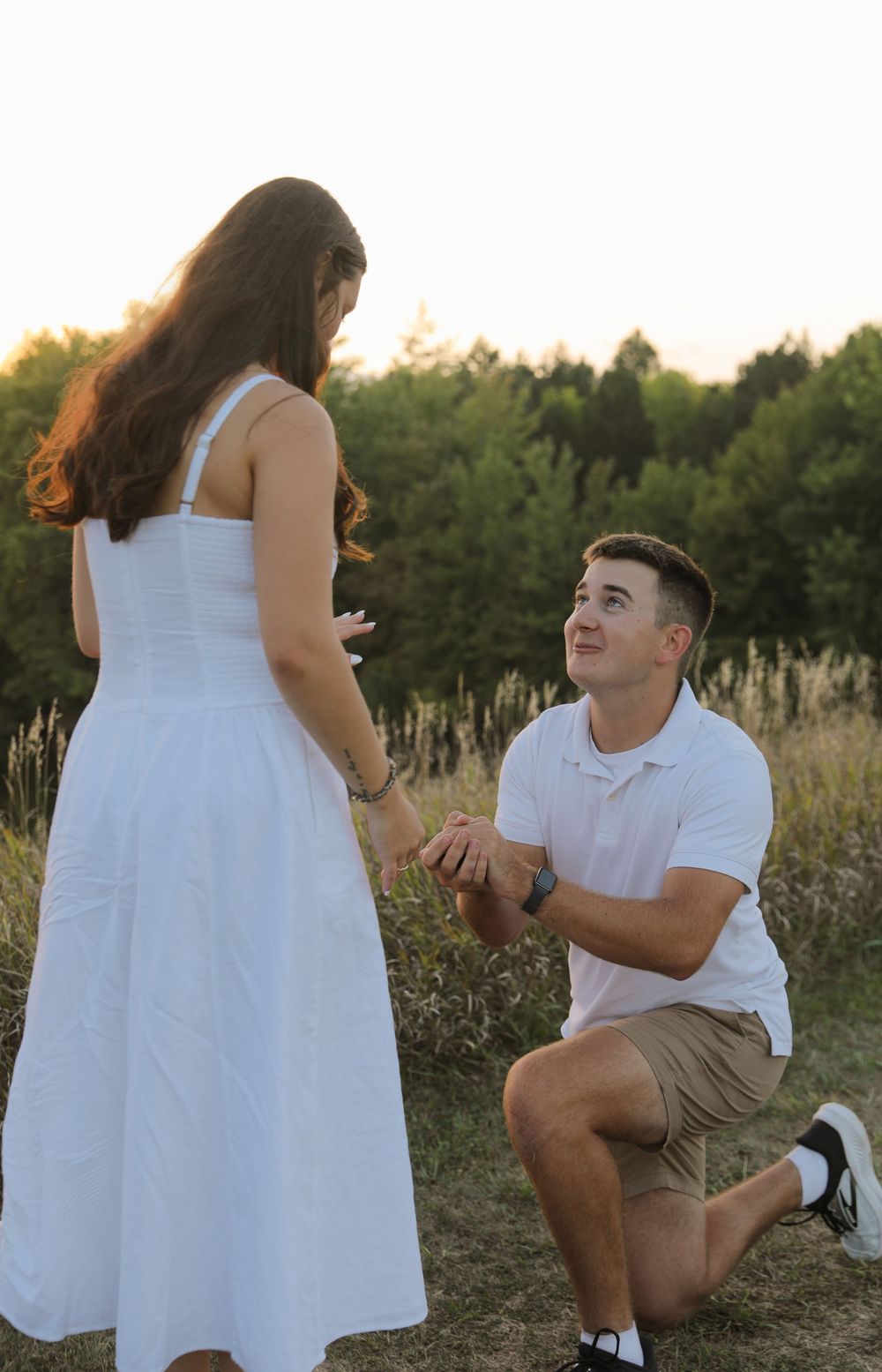 Man kneeling on one knee proposing to woman in white dress outdoors at golden hour in a field.