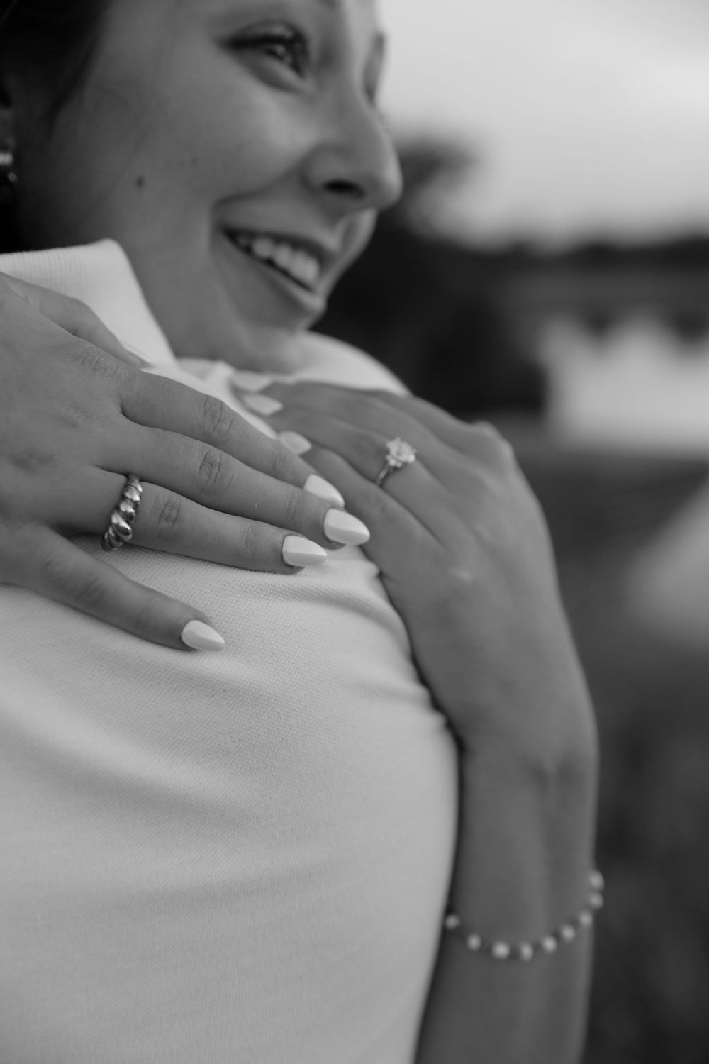 Black and white photo of a smiling bride showing her engagement ring and manicured nails on her shoulder.