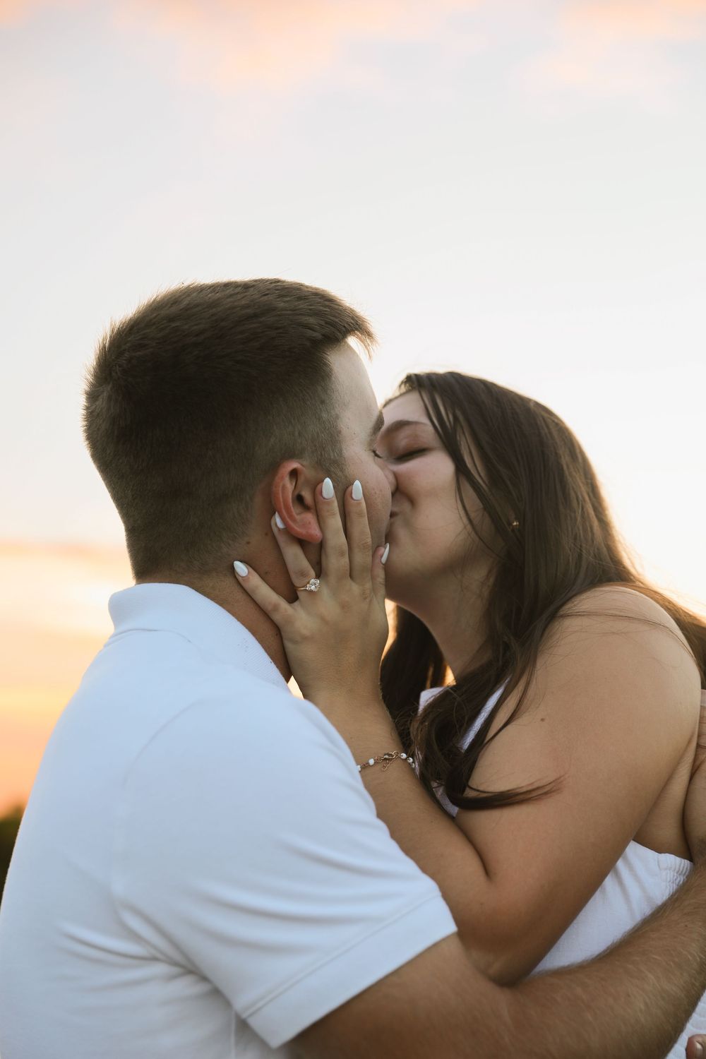 Couple sharing a romantic kiss at sunset, woman wearing engagement ring, golden hour sky in background.