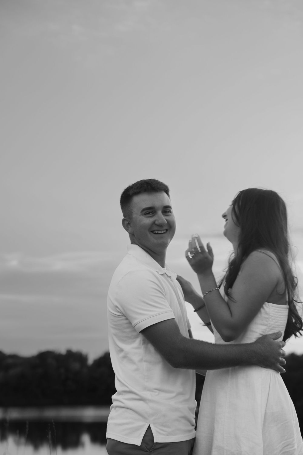 Black and white photo of a smiling couple embracing outdoors near a lake at sunset.