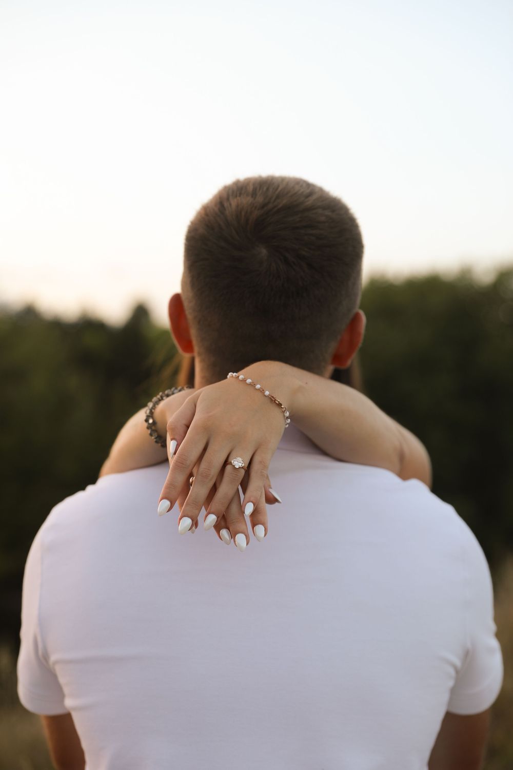 Couple embracing outdoors, woman's hand with engagement ring and bracelet resting on man's shoulder.