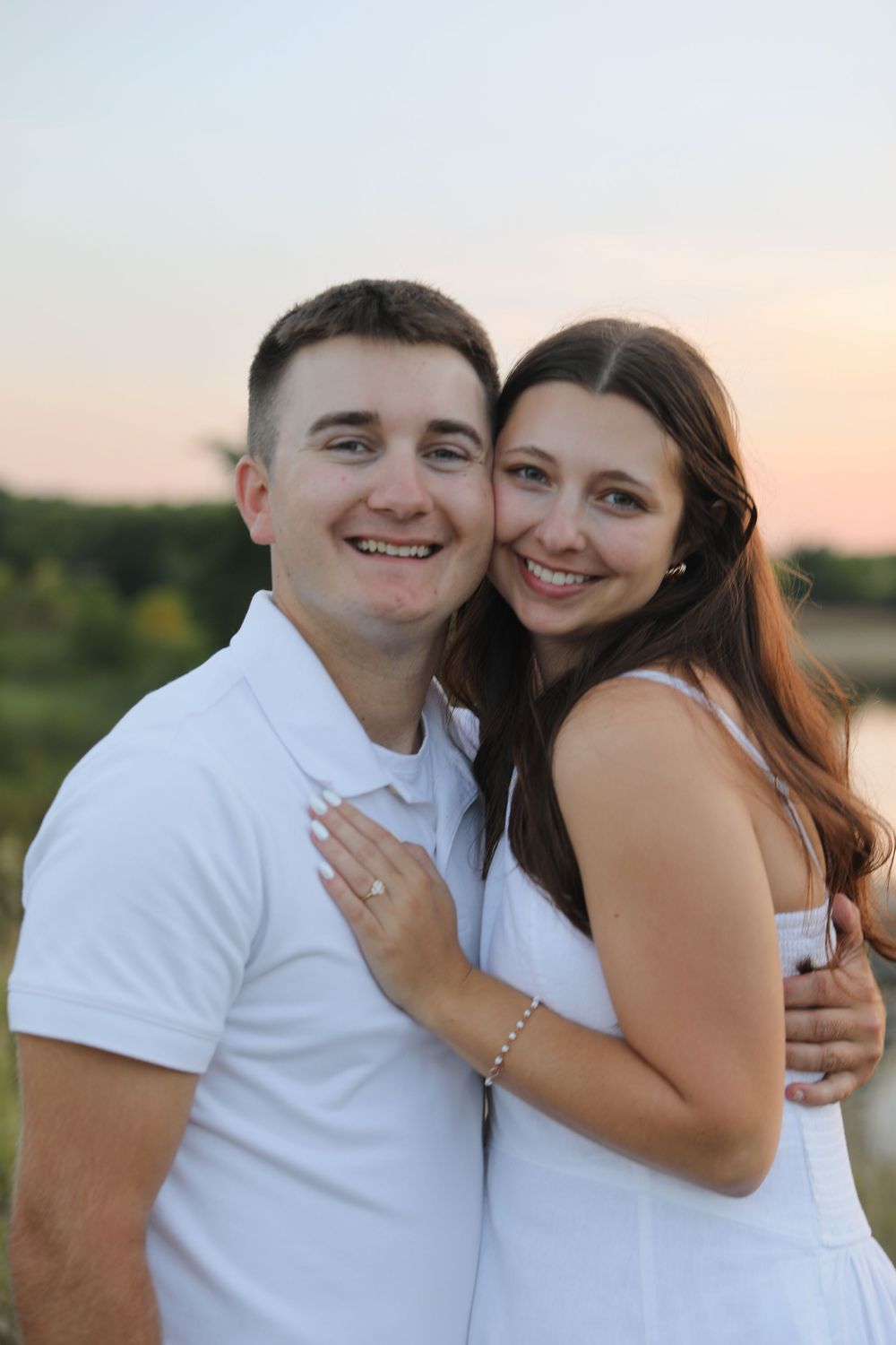 A smiling couple in white outfits embraces outdoors at sunset, with the woman showing an engagement ring.
