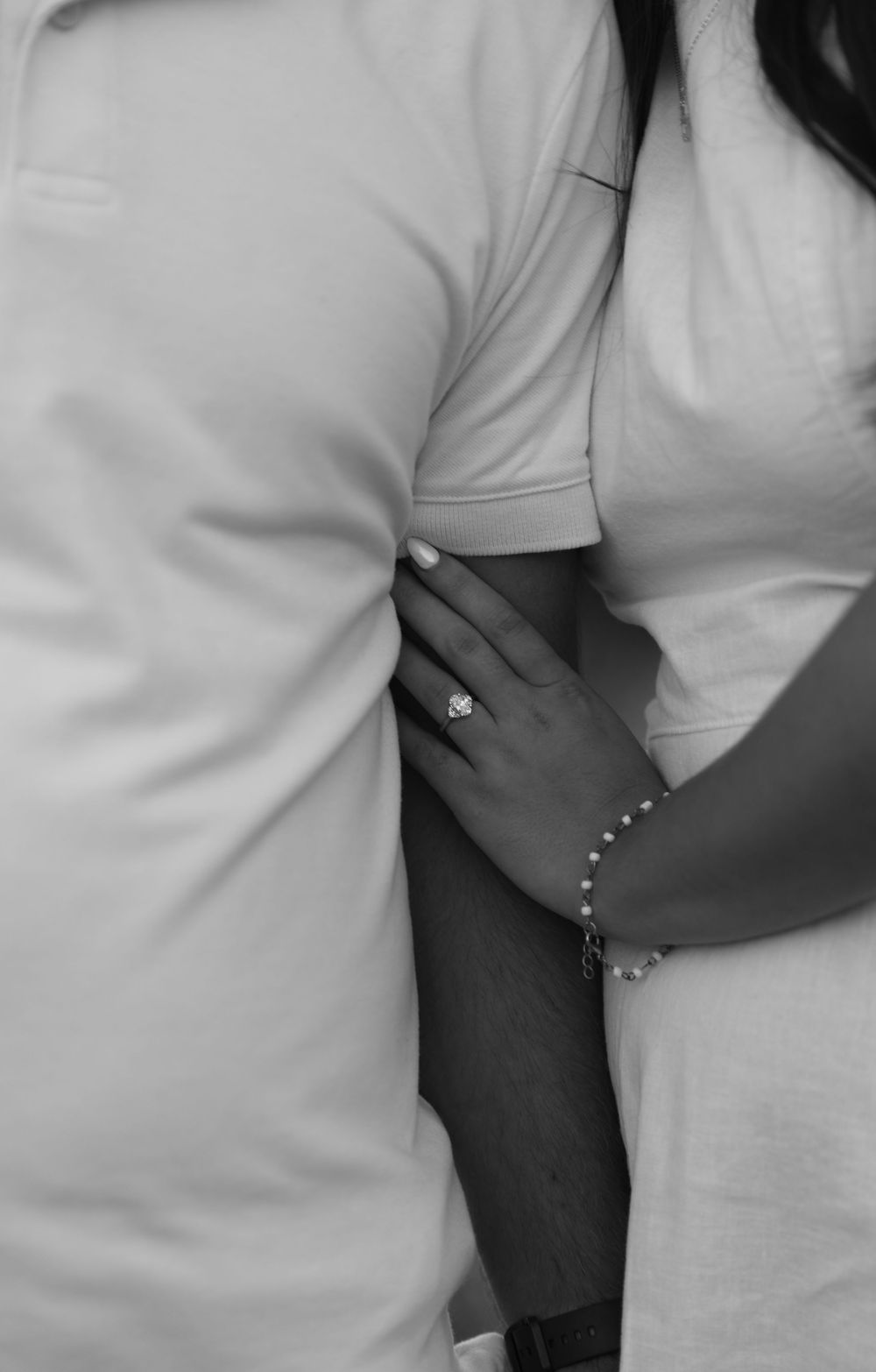 Black and white photo of a couple embracing, woman's hand showing an engagement ring and bracelet resting on man's arm.