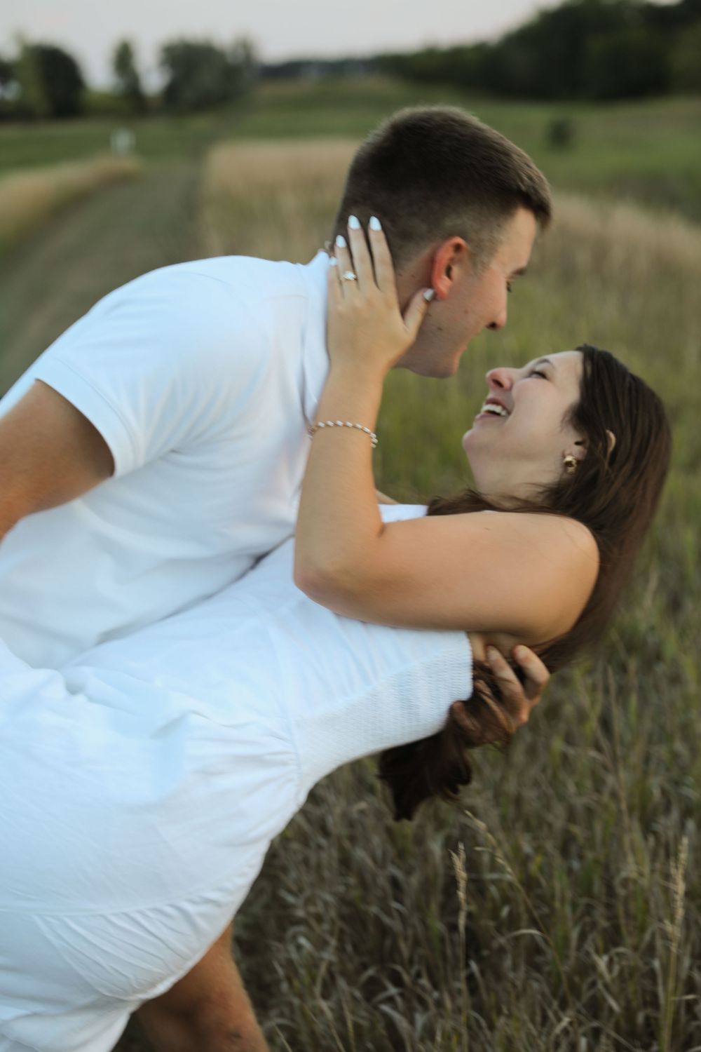 Couple in white outfits sharing a romantic moment in a green field, woman laughing as man dips her.