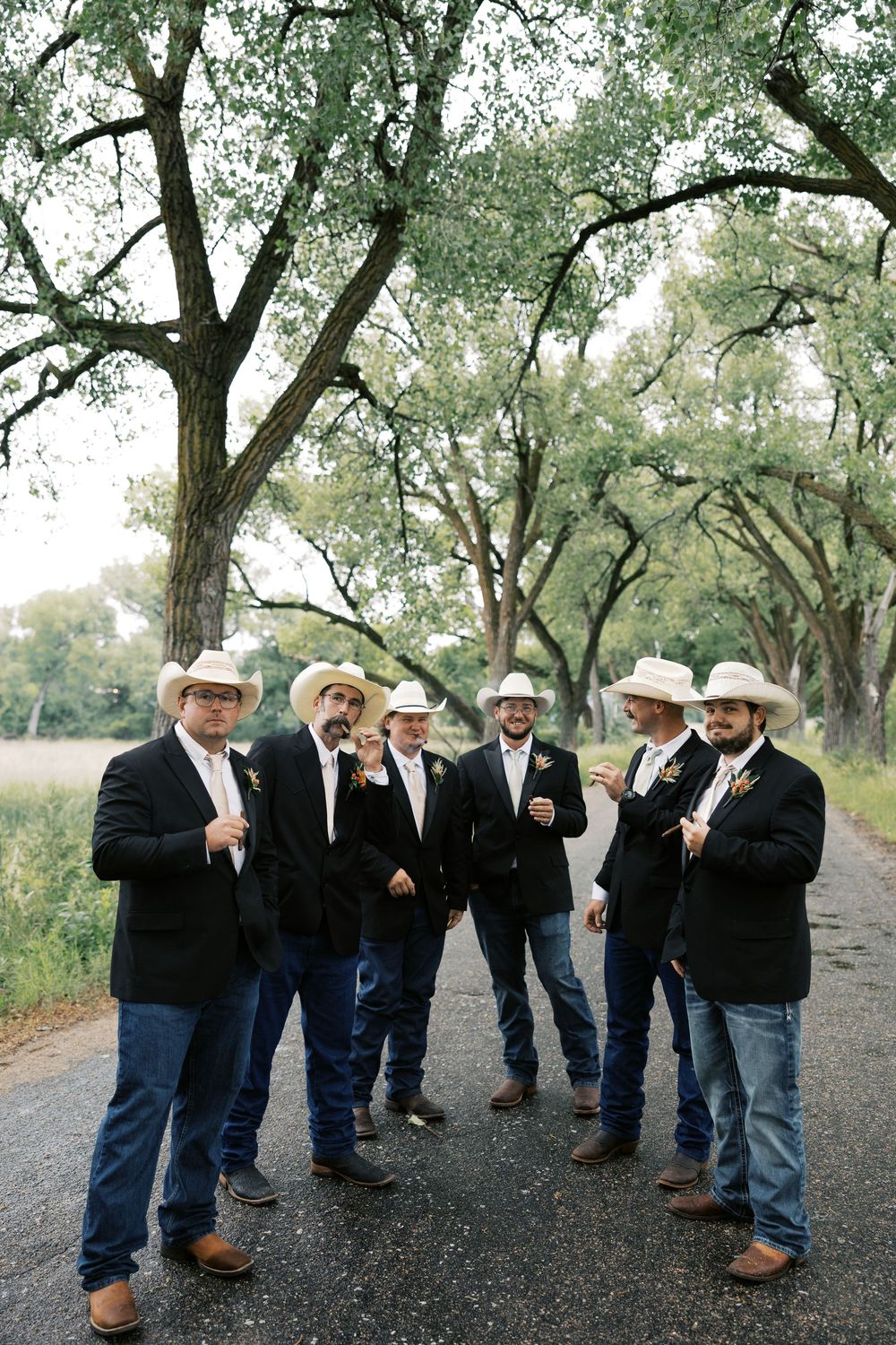 Groomsmen in black suits, cowboy hats, and boots pose on a tree-lined path at a rustic country wedding.